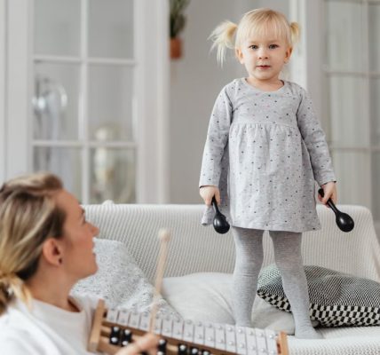 Young girl standing on couch holding percussion instruments, enjoying playful music time with adult nearby.