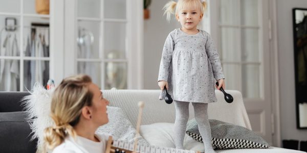 Young girl standing on couch holding percussion instruments, enjoying playful music time with adult nearby.