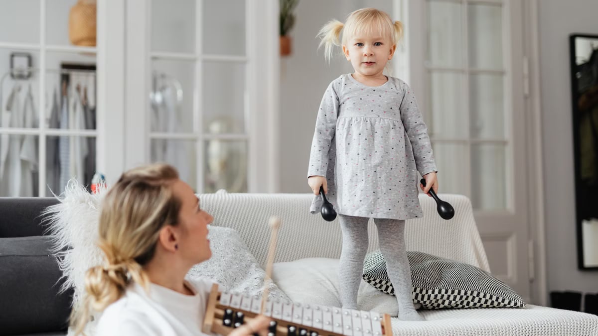 Young girl standing on couch holding percussion instruments, enjoying playful music time with adult nearby.